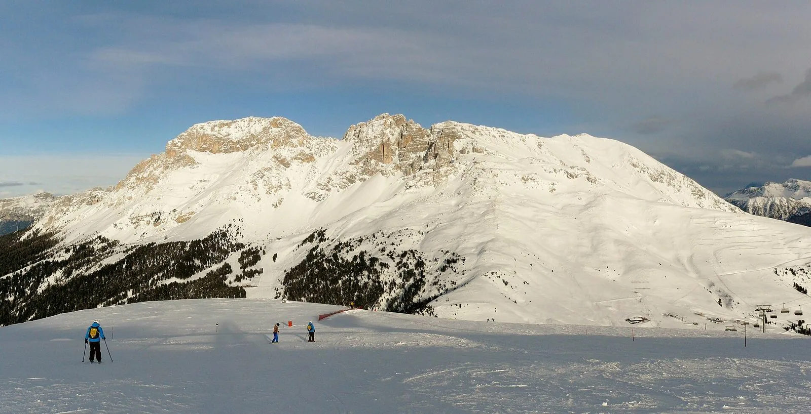 Il massiccio del Latemar innevato sotto un cielo velato, con boschi di conifere sulle pendici e, in primo piano, un'ampia pista battuta con alcuni sciatori in lontananza