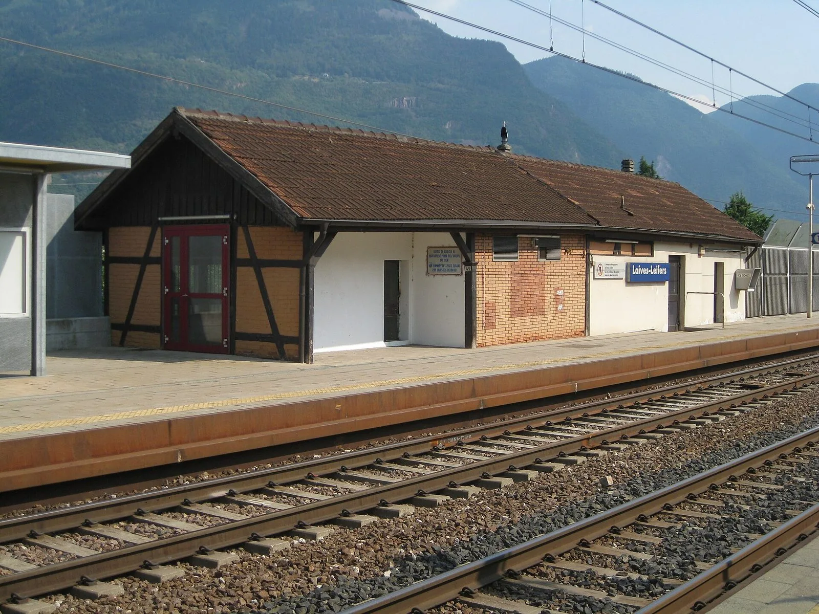 La piccola stazione ferroviaria di Laives-Leifers: un edificio basso con tetto in coppi e parte a graticcio, il marciapiede con la scritta blu del nome bilingue, i binari in primo piano e i pendii boscosi della valle sullo sfondo