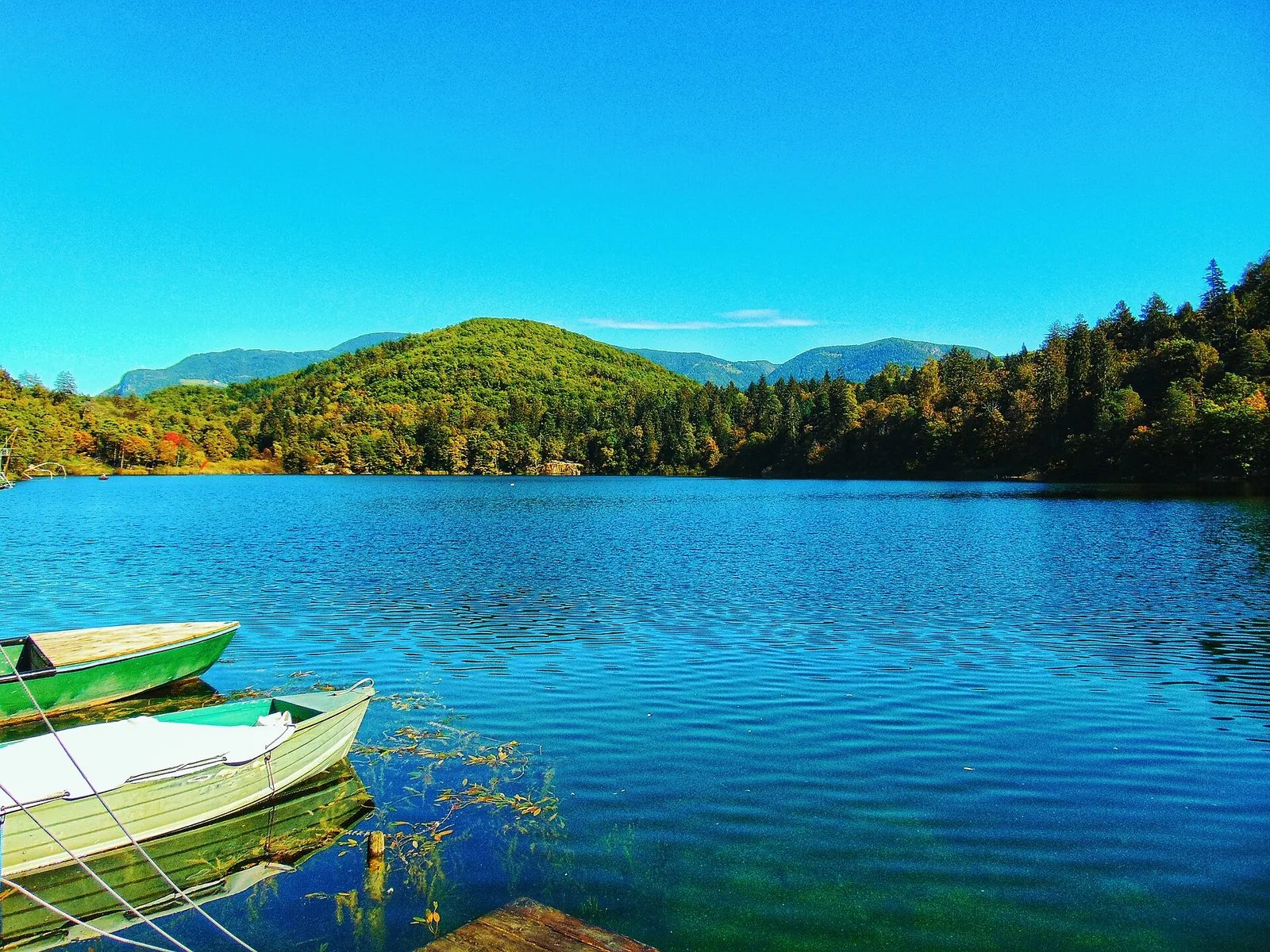 Il Lago Grande di Monticolo in una giornata limpida: acqua blu increspata, due barche a remi ormeggiate a un pontile di legno in primo piano e rive coperte di bosco, con montagne sullo sfondo