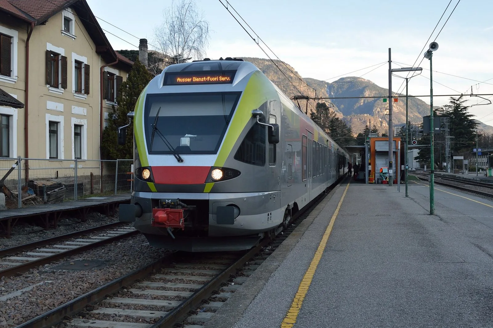 Un treno regionale altoatesino a pianale ribassato, grigio con fasce verde chiaro e rosse, fermo al binario di una stazione con un edificio storico color crema a sinistra e montagne rocciose illuminate dal sole sullo sfondo
