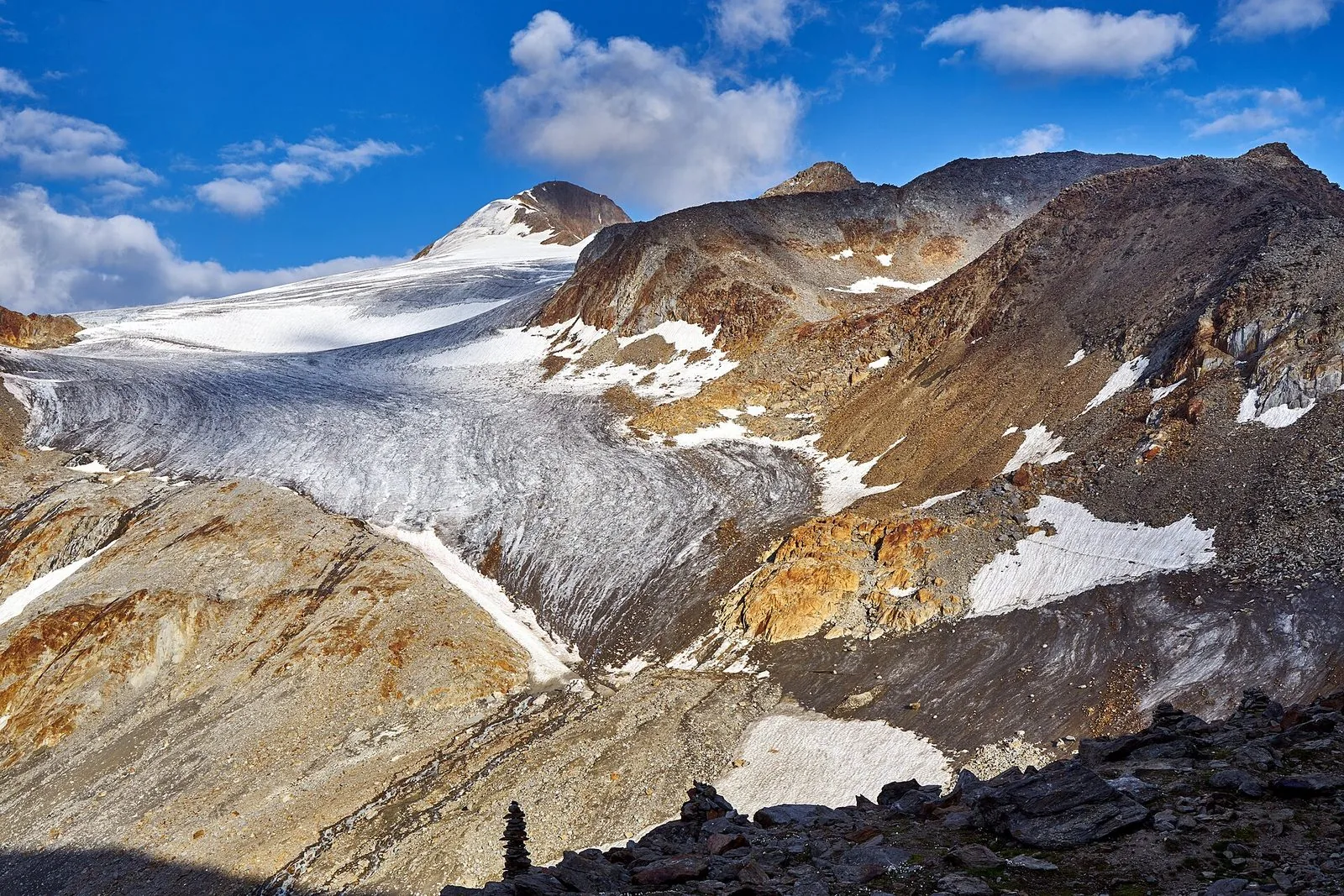 Il ghiacciaio del Similaun nelle Alpi Venoste: una lingua di ghiaccio grigio e bianco scende fra pendii rocciosi dai toni ocra e rossicci, con la cima innevata sotto un cielo azzurro con nuvole bianche e un piccolo ometto di pietre in primo piano